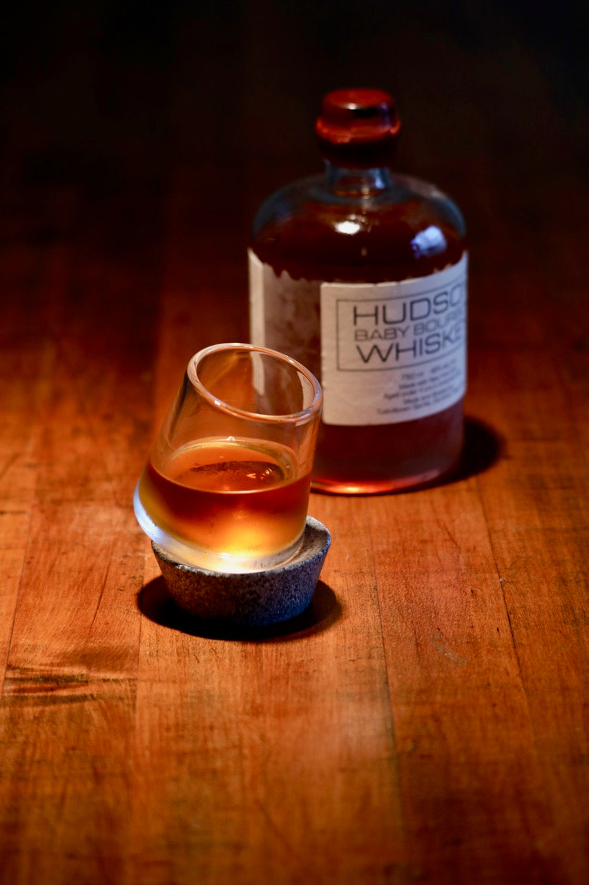Glass of whiskey on a gray soapstone base with whiskey bottle in background on wooden surface.