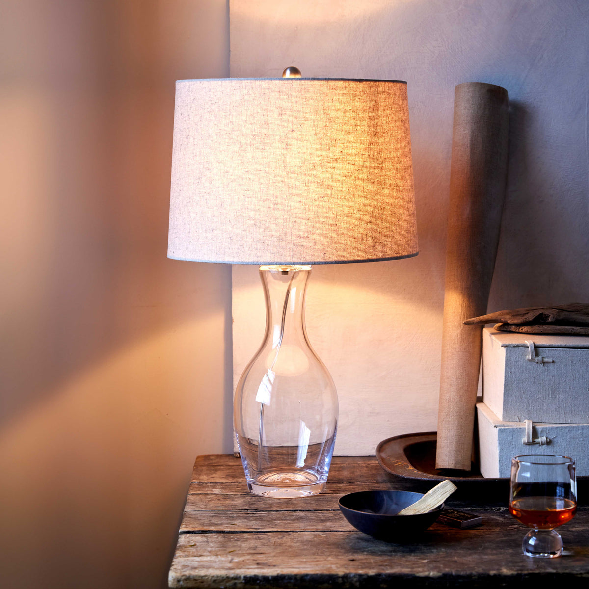 Table lamp with clear glass bottle base, linen shade, and brass finial on wooden surface.