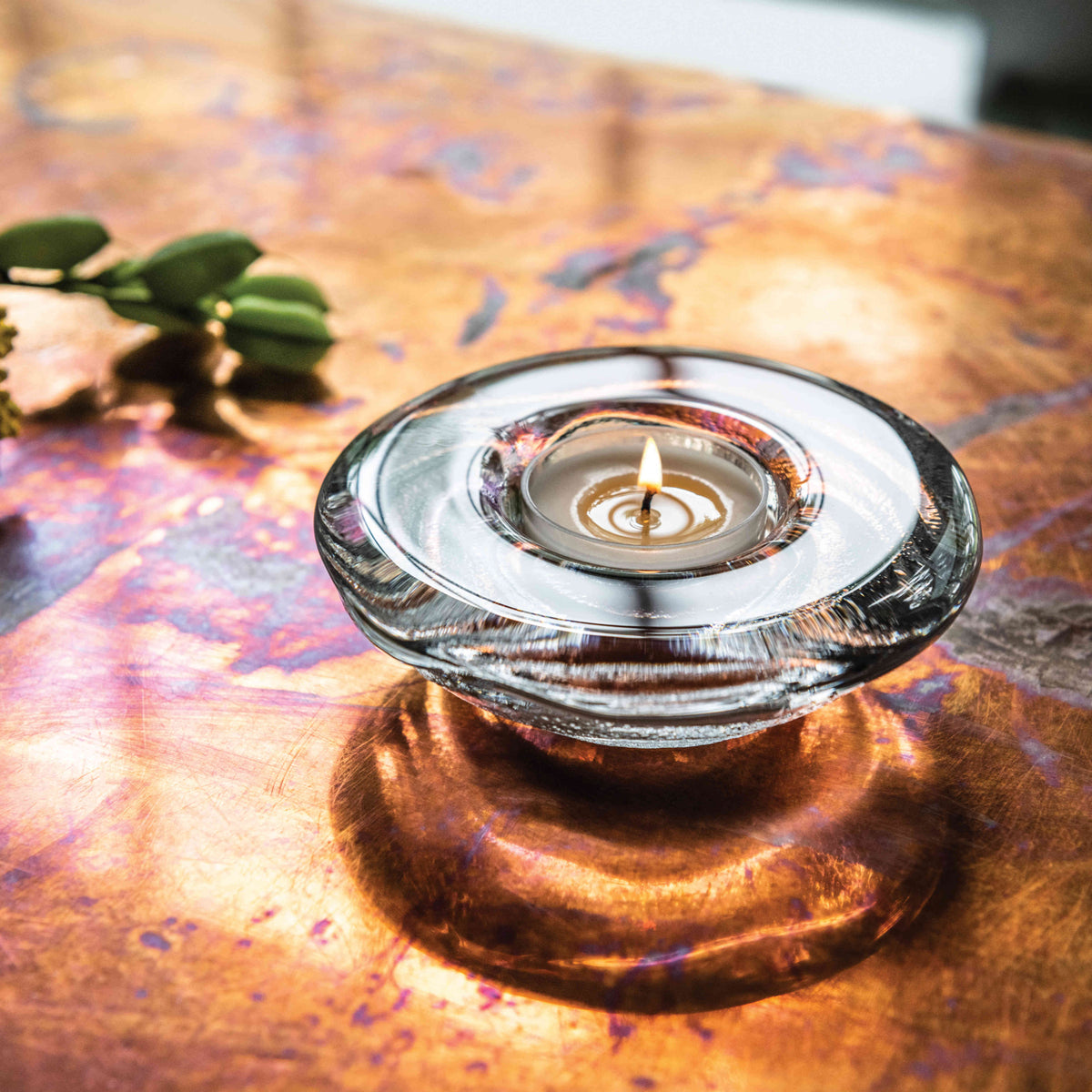 Glass tealight holder with lit candle on patterned surface beside greenery.