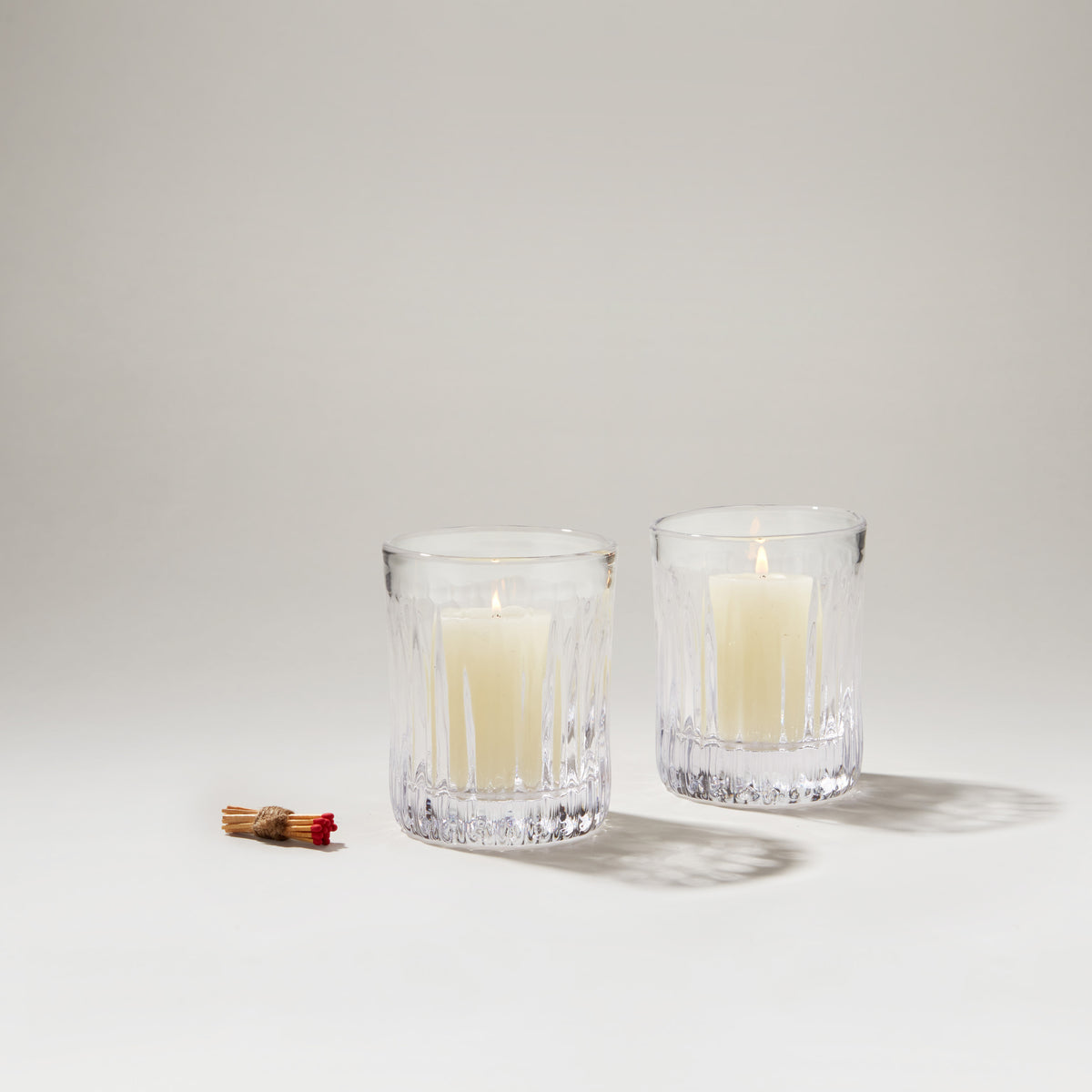 Two clear textured glass hurricane holders with lit cream candles inside and decorative twine bundle beside them.