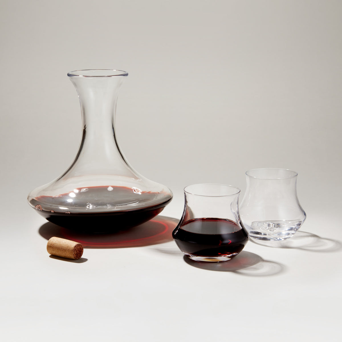 Glass decanter filled with red wine, two wine glasses, and a cork on neutral background.