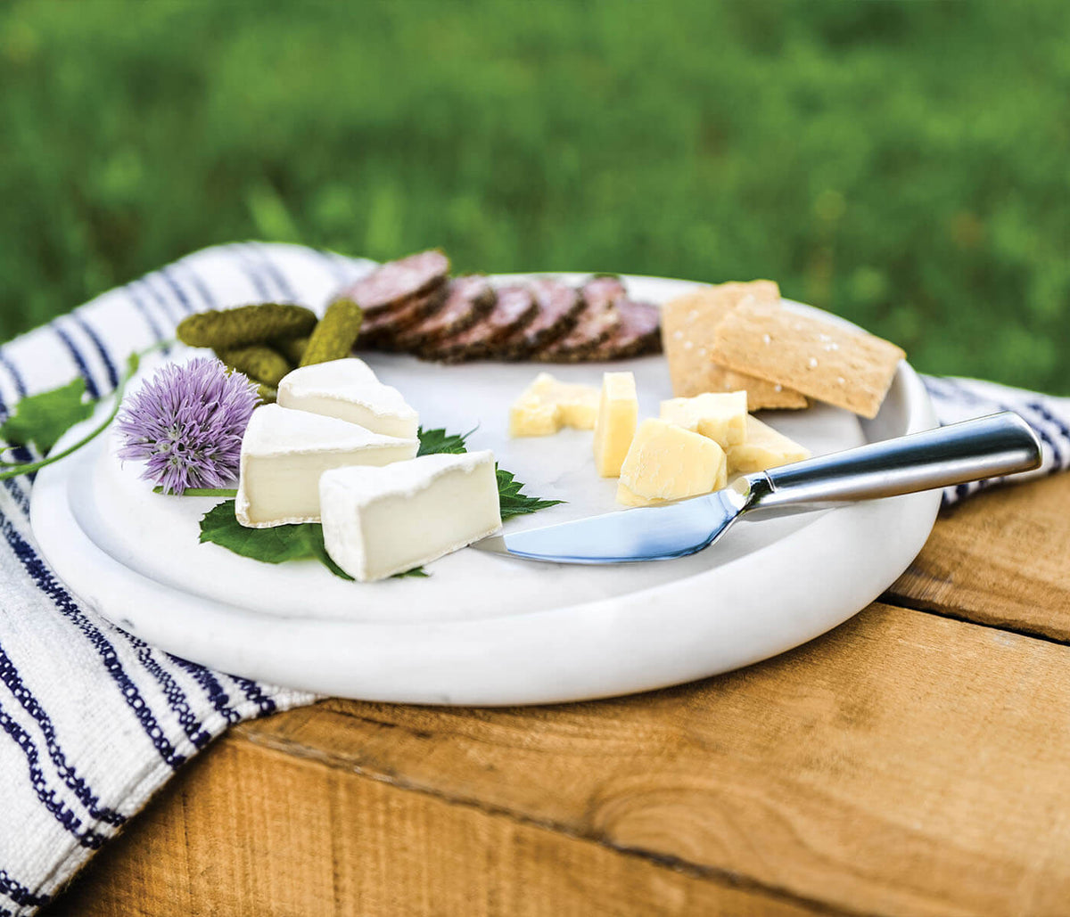 White marble round cheese board with assorted cheeses, crackers, pickles, and a cheese knife on a wooden surface.