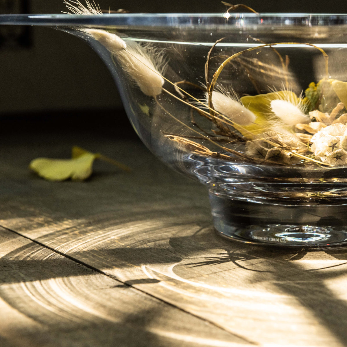 Clear glass bowl filled with dried grasses, wheat, and white botanicals, displayed on wooden surface.