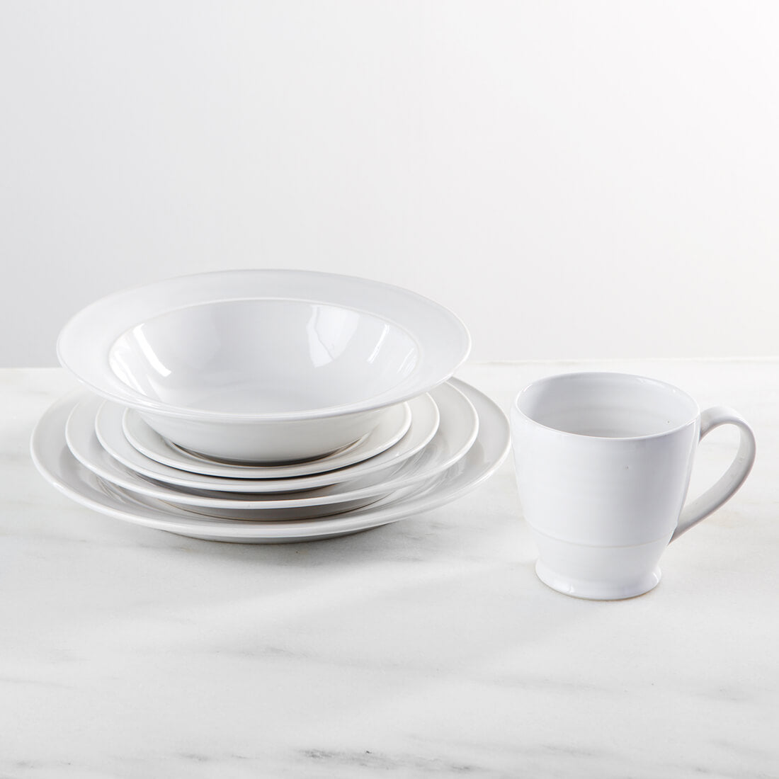 White dinnerware set with stacked plates in graduated sizes, a bowl, and a mug displayed on marble surface.