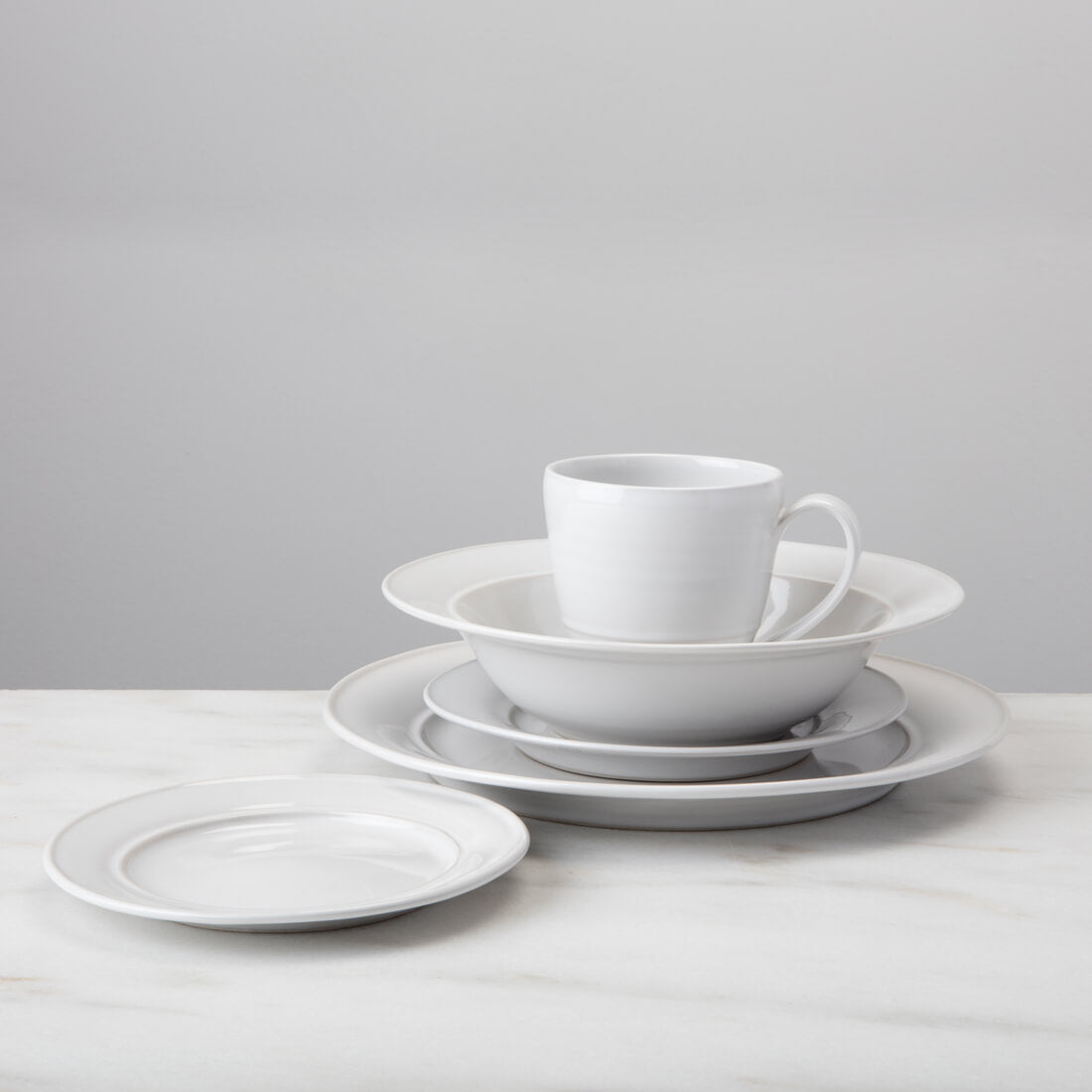White Cavendish dinnerware set displayed with stacked plates, bowls, cup, and saucer on marble surface.