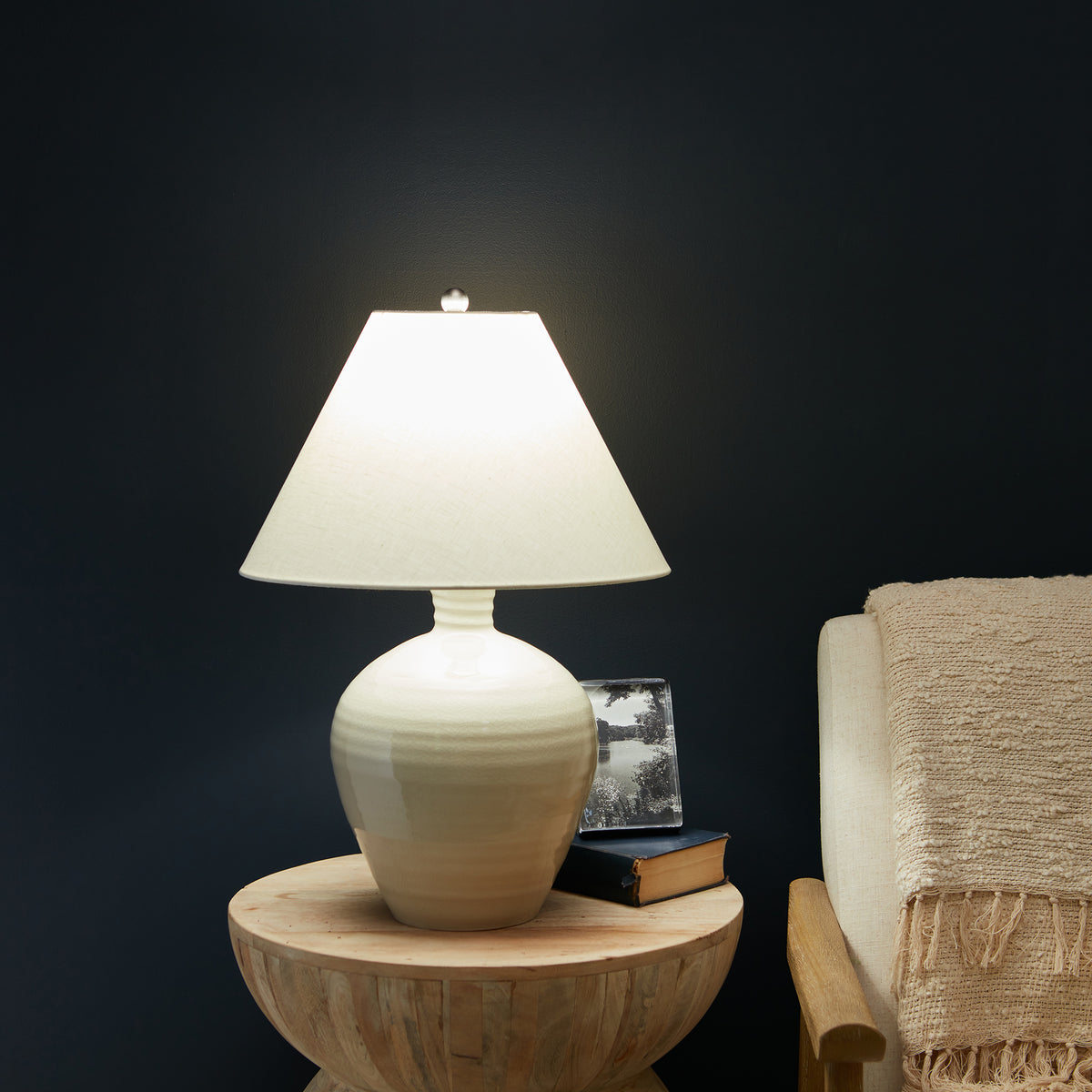 Cream pottery lamp with rounded bulbous base and tapered white linen shade on wooden table.