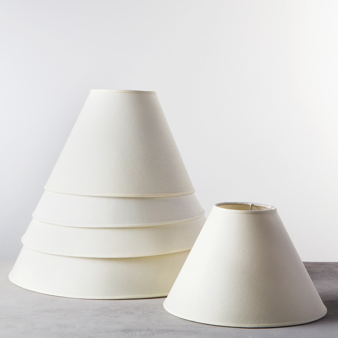 Cream linen empire lampshades in two sizes, displayed side by side on gray surface.