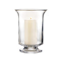 Clear glass hurricane vase with pedestal base holding a lit cream-colored pillar candle.