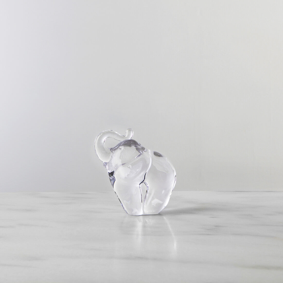 Clear glass elephant figurine with raised trunk on a marble surface against a gray background.