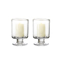 Two clear glass hurricane candle holders with pedestal bases, each containing a cream-colored pillar candle.