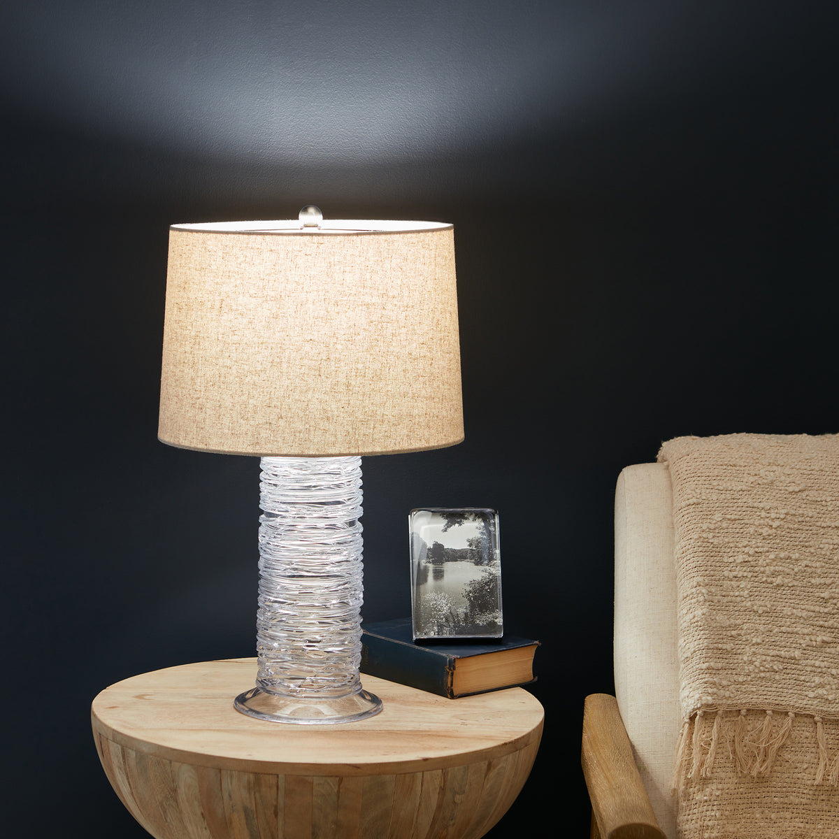 Table lamp with wrapped white base, beige linen shade, on wooden round table next to books and framed photo.
