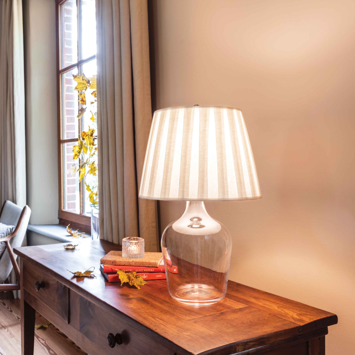 Table lamp with clear glass base and beige striped fabric shade, displayed on wooden desk.