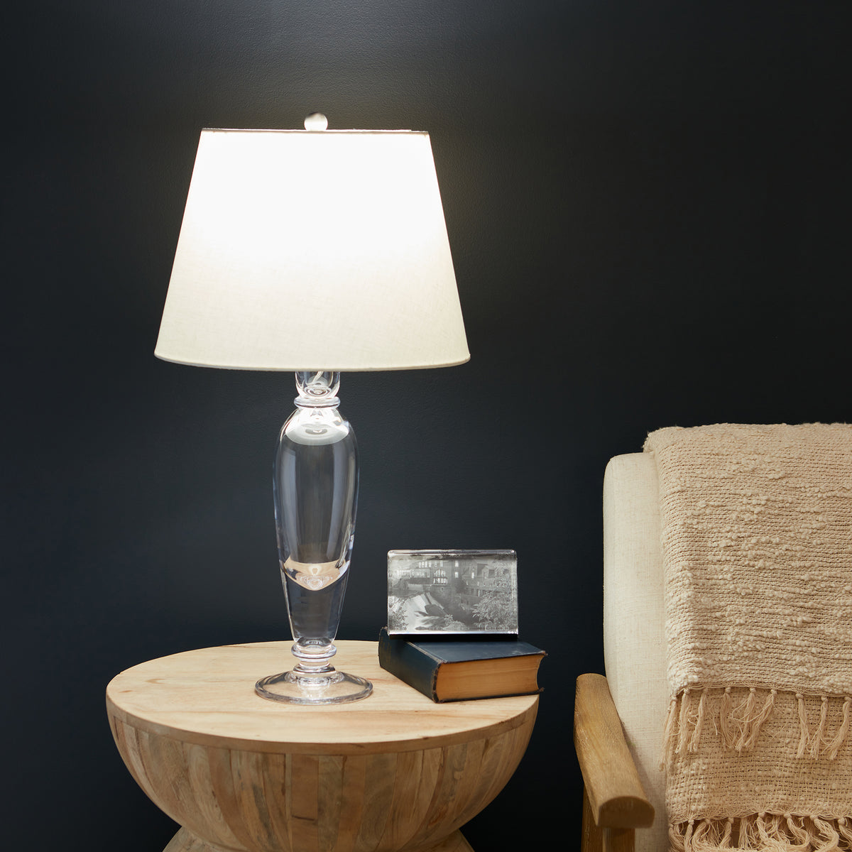 Clear glass table lamp with cream fabric shade on wooden round side table next to upholstered chair.