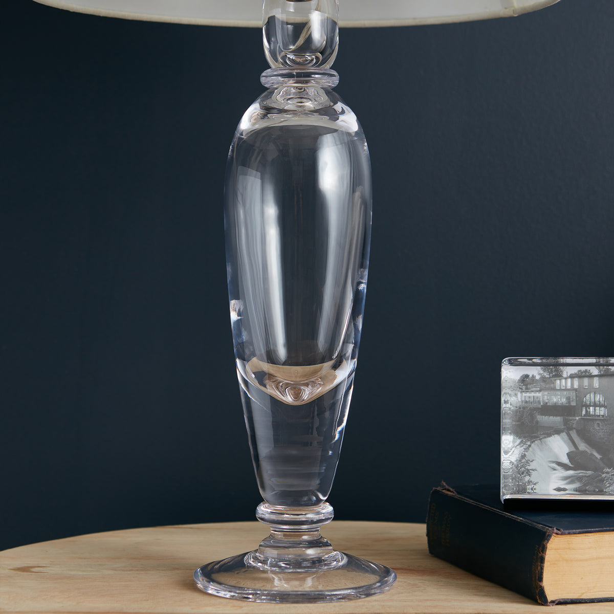 Clear glass table lamp with elongated cylindrical body, decorative band detail, and cream linen shade on dark blue background