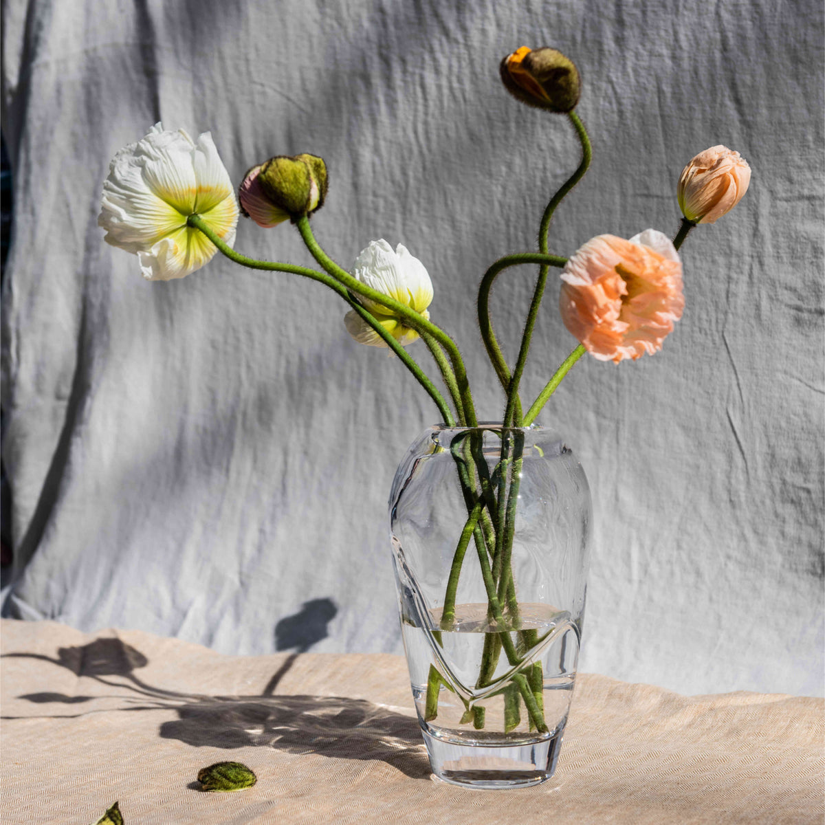 Clear glass vase with white and peach flowers and green stems arranged in water against a gray linen backdrop.