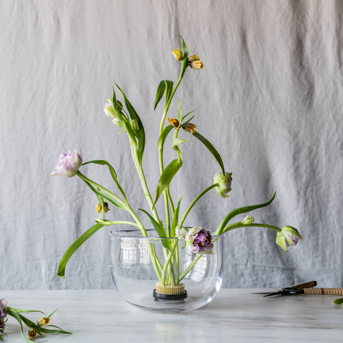 Clear glass bowl with grid pattern containing fresh flowers in various stages of bloom with green stems and leaves.