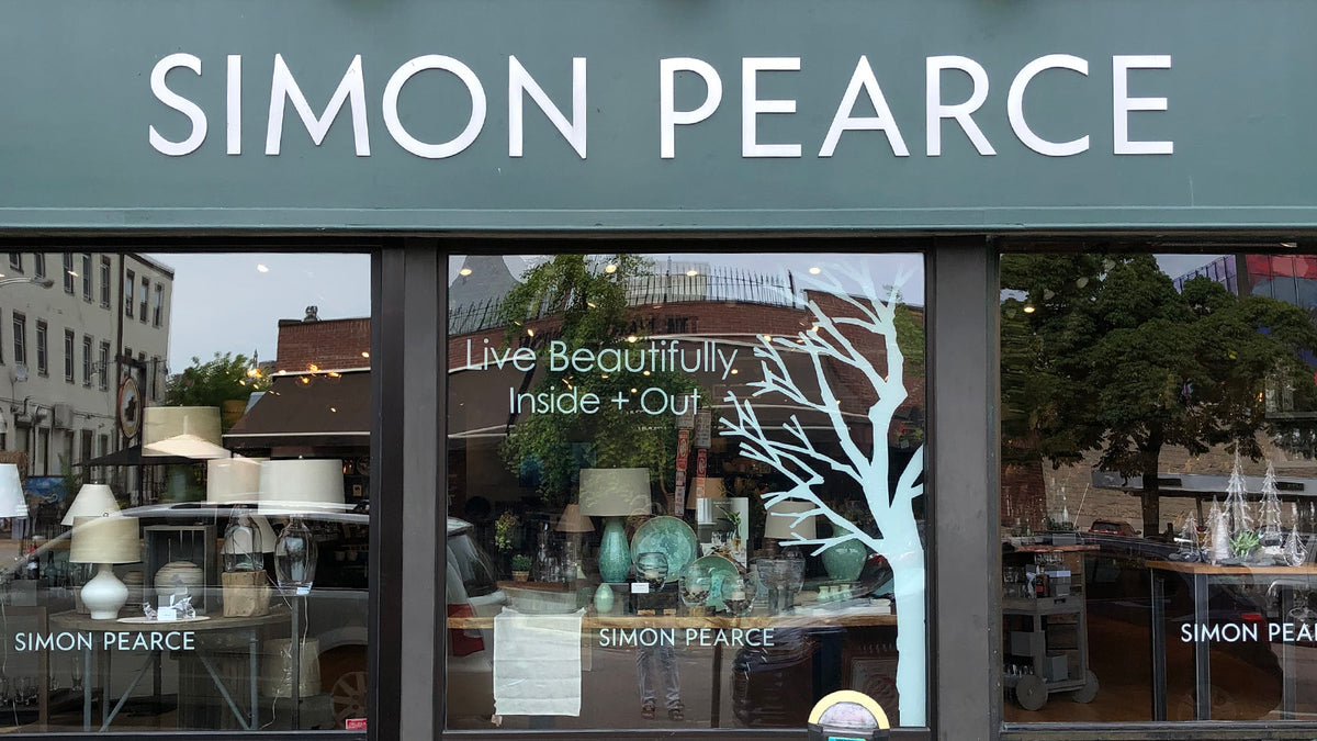 Exterior of the Simon Pearce retail store in Burlington, Vermont, with a teal fascia sign and large display windows featuring lamps, glassware, and the tagline 'Live Beautifully Inside + Out'