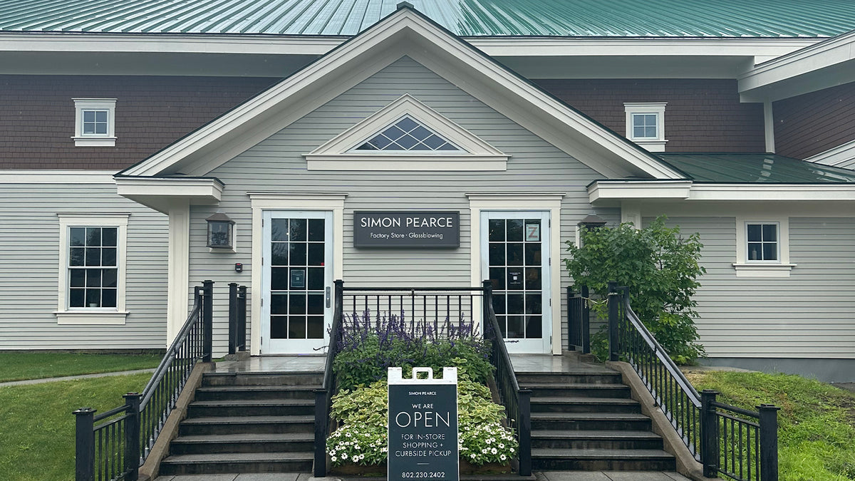 Exterior of the Simon Pearce Factory Store and Glassblowing and Pottery Workshop in Windsor, Vermont, a gray clapboard building with a green metal roof, black railings, and an 'Open' sandwich board sign at the entrance