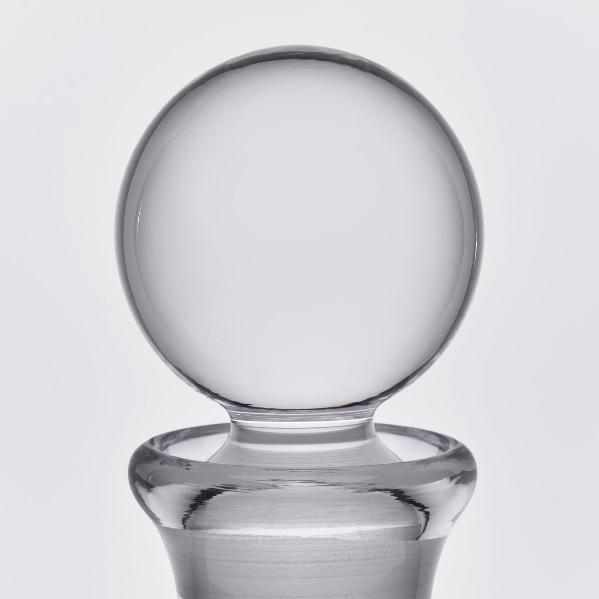 Clear glass decanter with spherical stopper and wide base on neutral background.