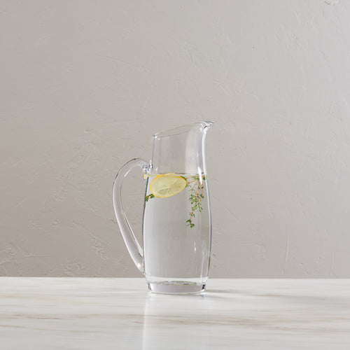 Clear glass pitcher with handle containing water, lemon slice, and fresh herbs.