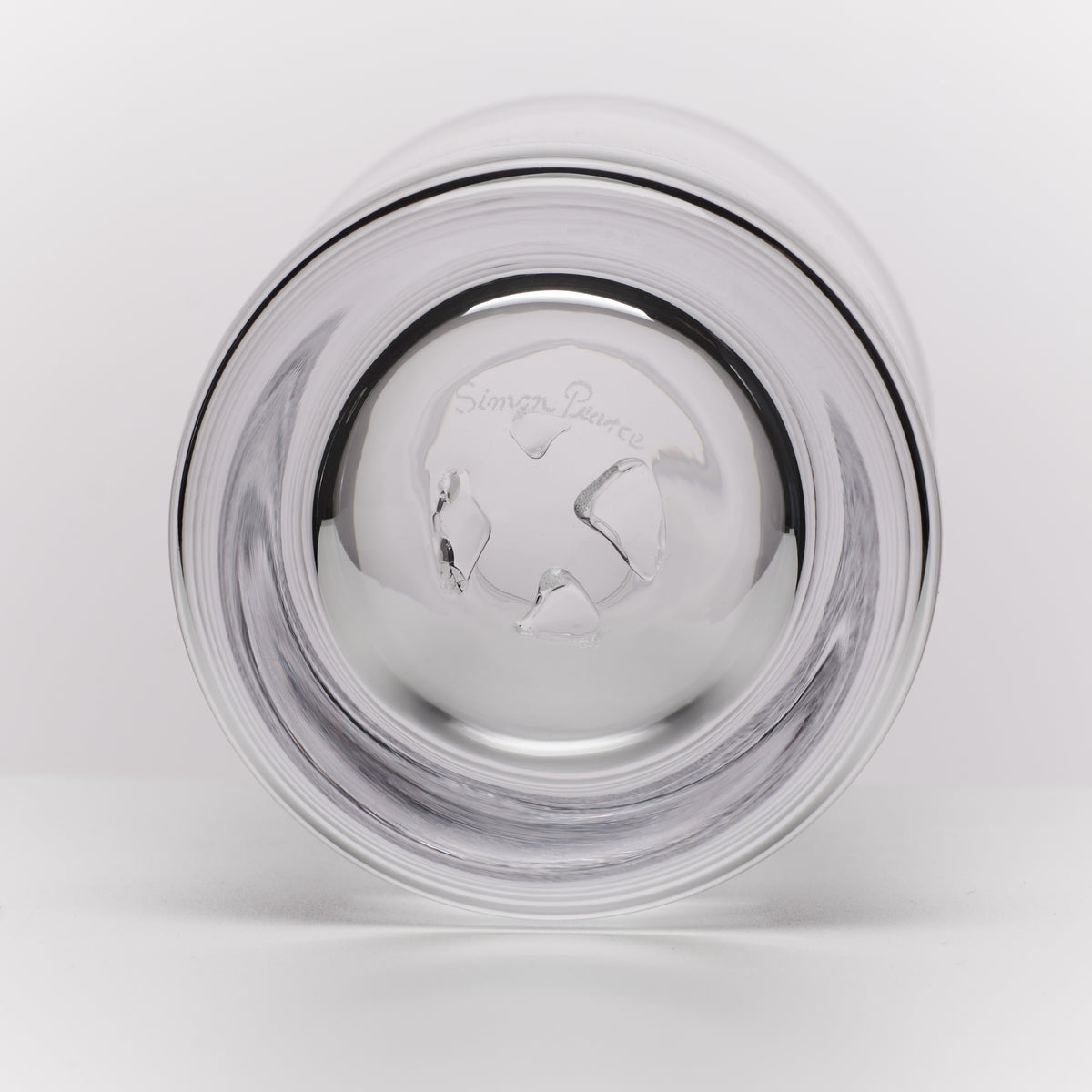 A clear glass tumbler shown from the bottom on a light gray background. The brand name 'Simon Pearce' is engraved on the bottom of the glass.