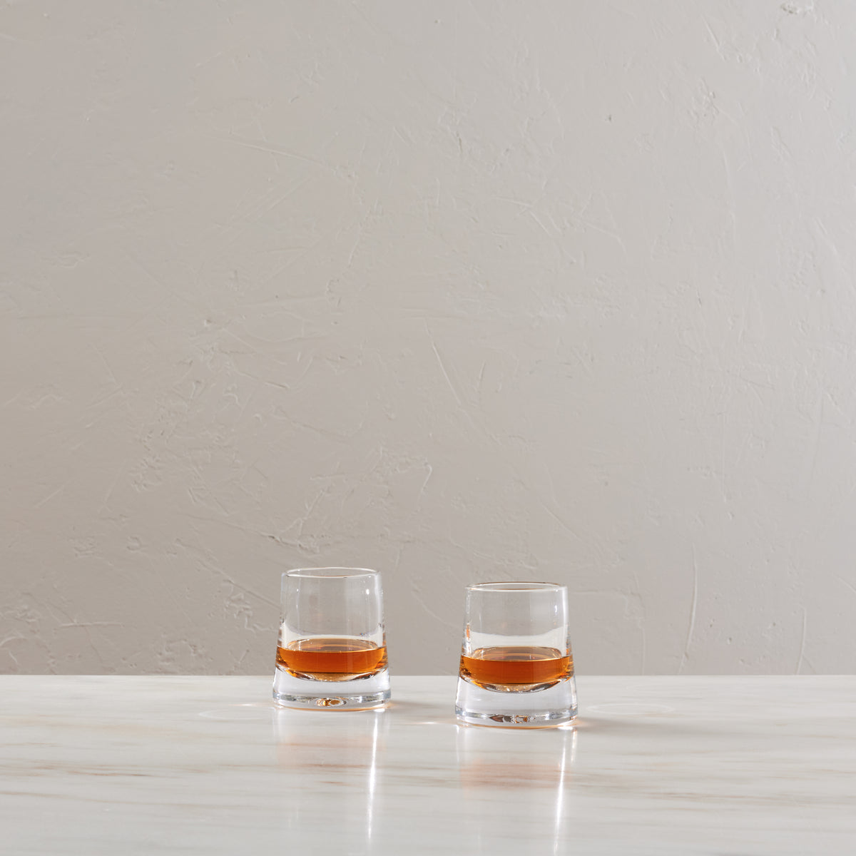 Two glasses filled with amber bourbon liquid on a white surface against a beige textured wall.