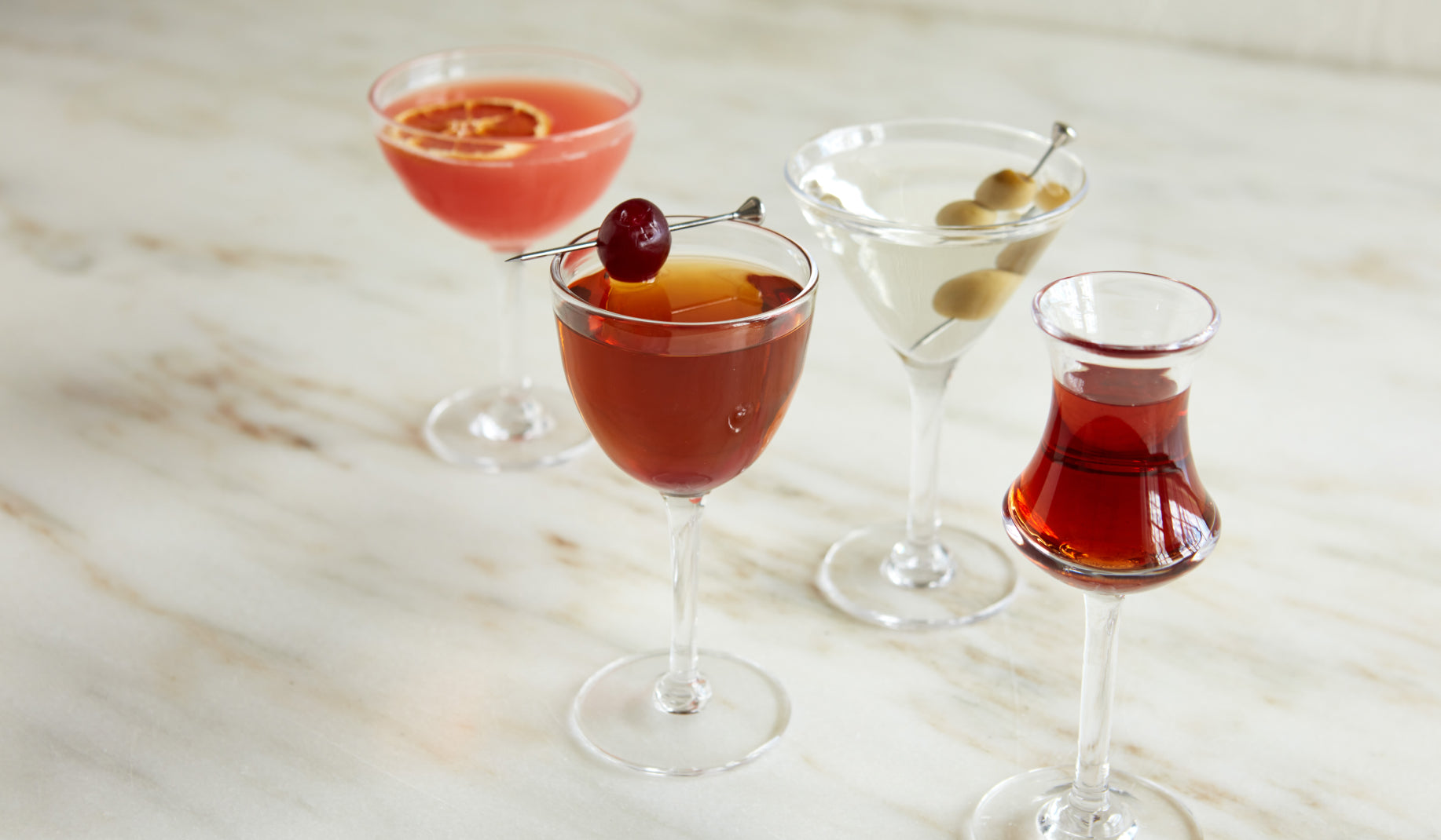Four Simon Pearce Dorset cocktail glasses filled with assorted garnished drinks arranged on a marble surface
