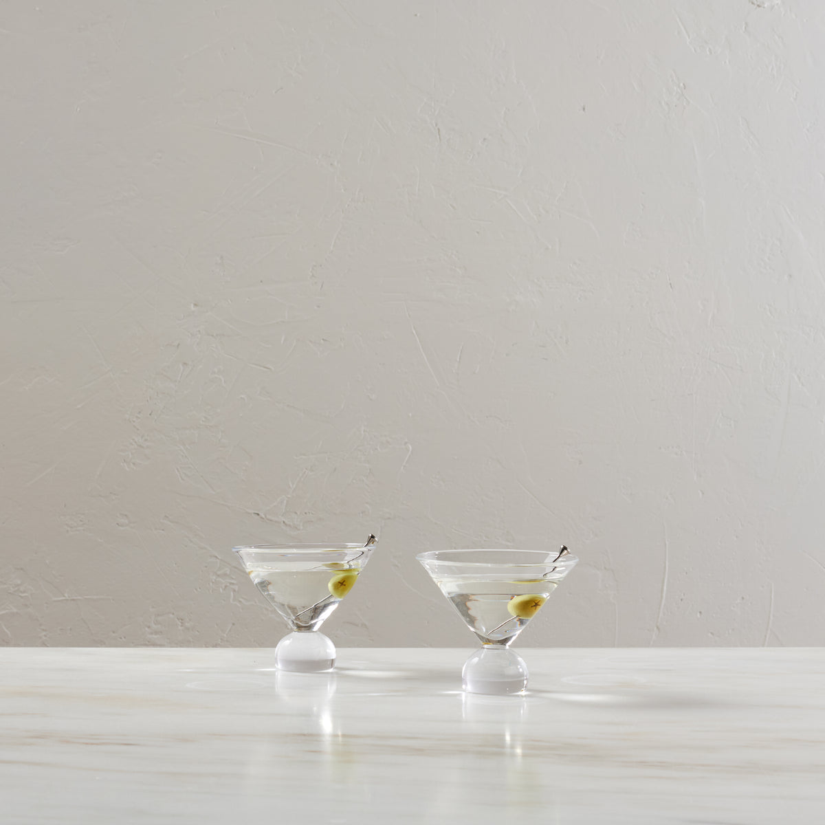 Two martini glasses filled with clear liquid and green olives on white pedestals against a textured beige wall.