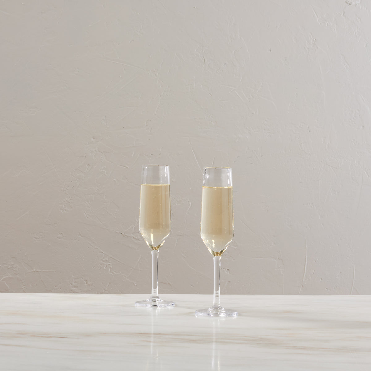 Two clear champagne flutes filled with light-colored liquid on a white surface against a textured beige wall.