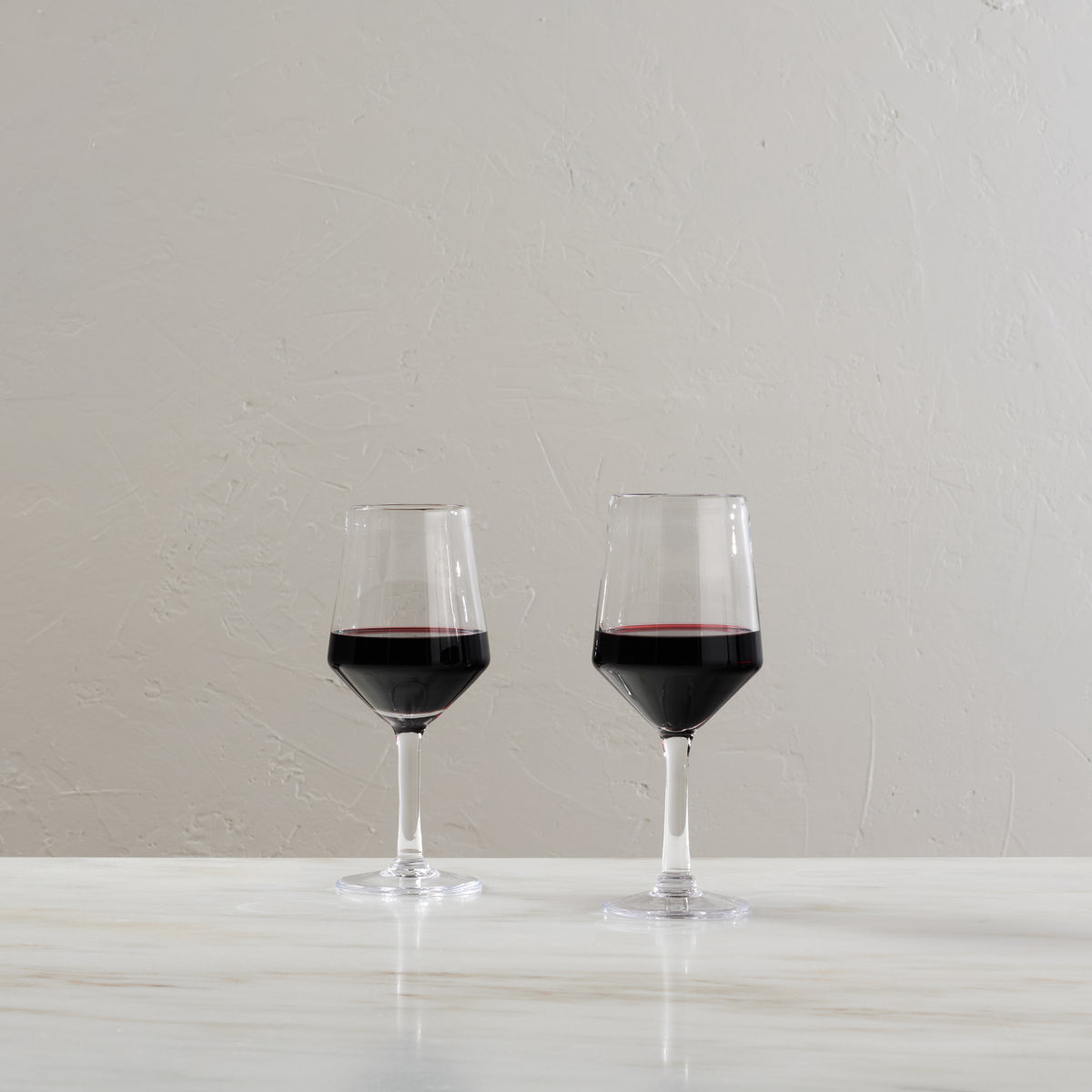 Two wine glasses filled with red wine on a white surface against a textured gray wall.