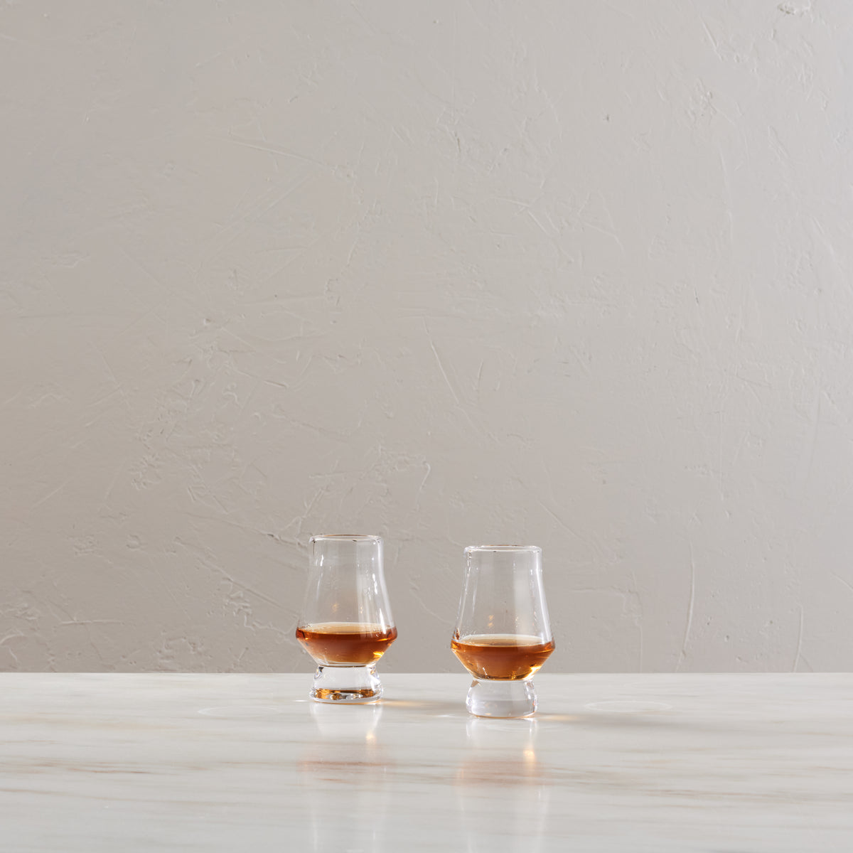 Two clear tasting glasses with tulip-shaped bowls containing amber liquid, displayed on a white surface.