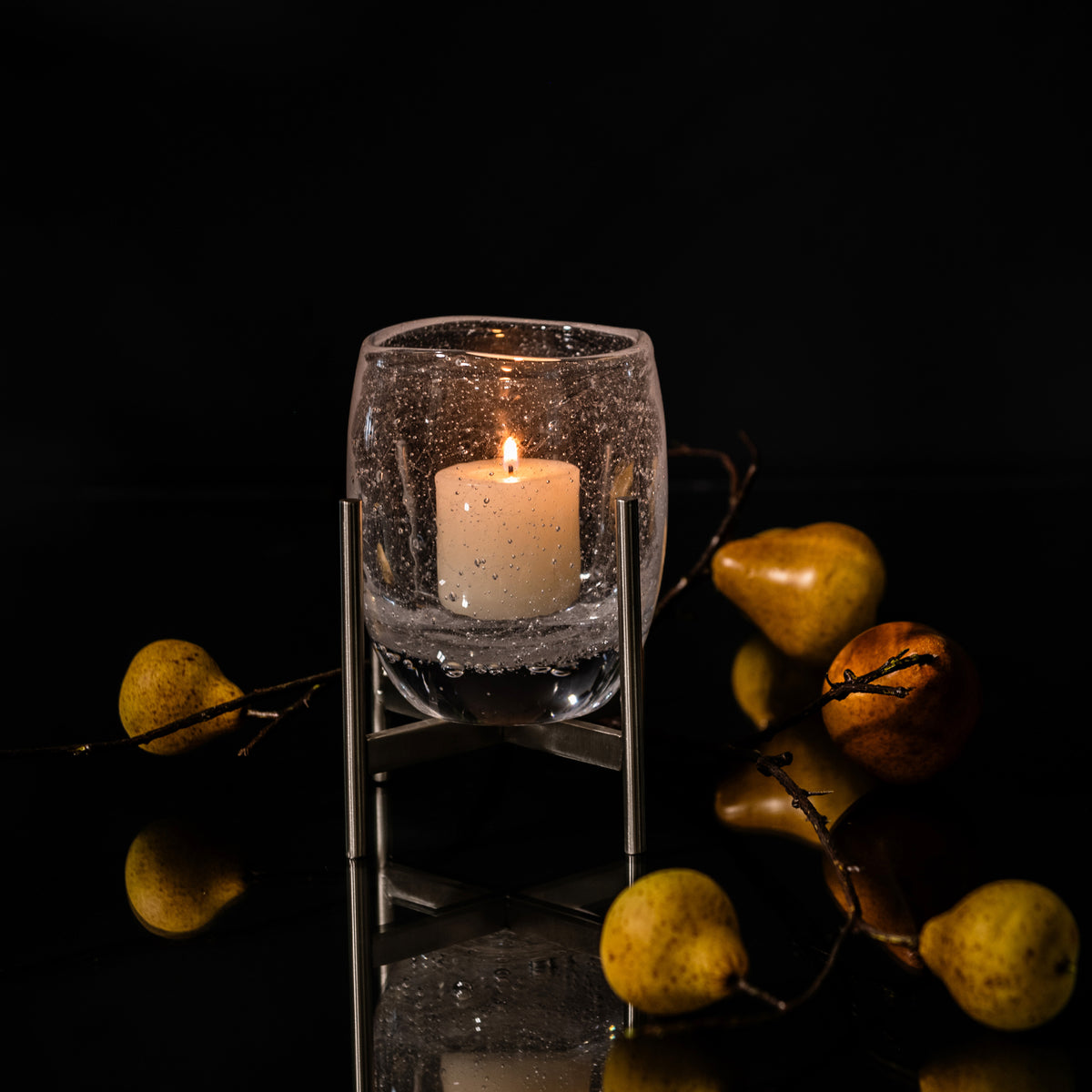 Textured glass hurricane with lit cream candle inside, mounted on stainless steel stand, surrounded by decorative pears and b