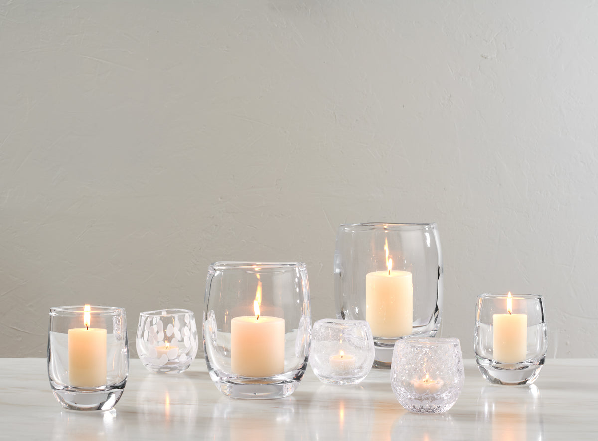 Set of six clear glass hurricane votive candle holders in varying sizes with lit cream-colored candles inside.