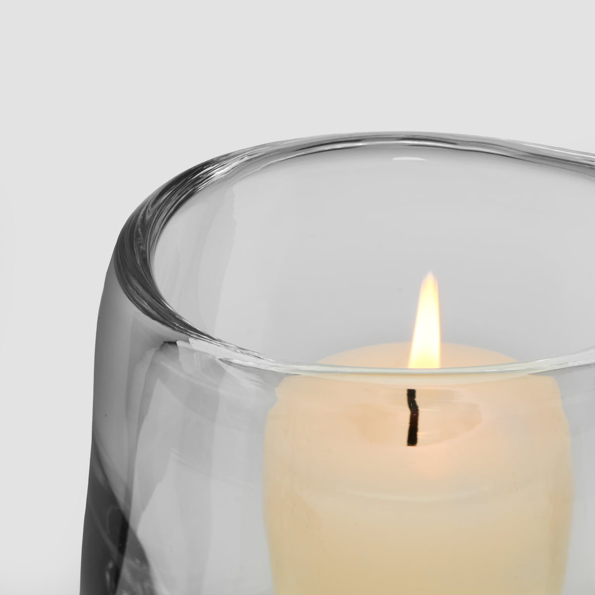 Cream-colored lit candle in clear glass hurricane holder with black wick and protective wrapping.