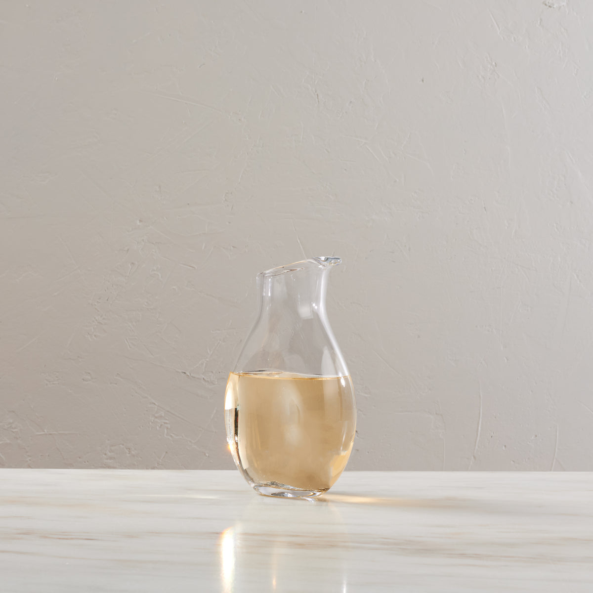 Clear glass carafe with curved body filled with pale liquid, pouring spout at top right, positioned on white surface.