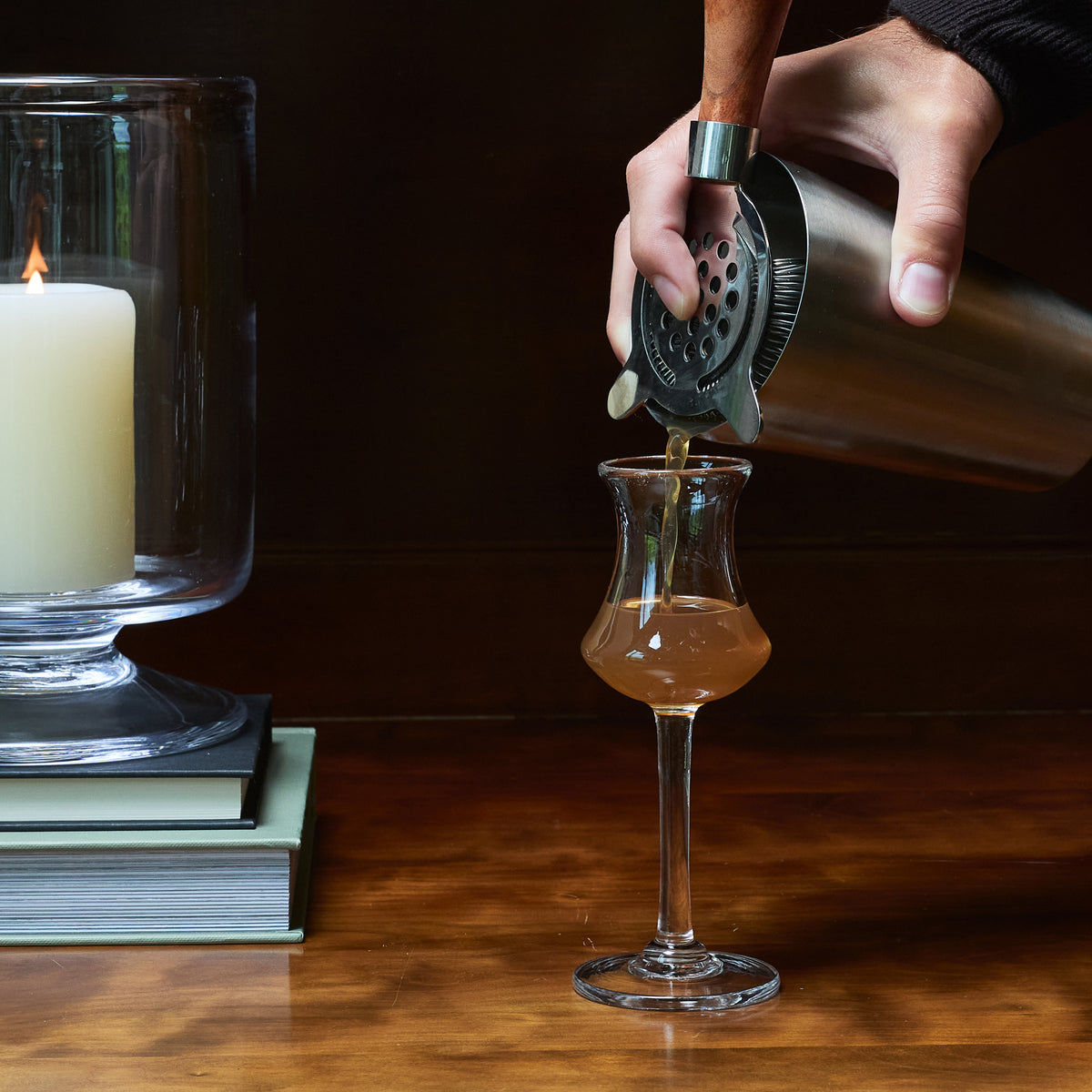Hand pouring amber liquid from a cocktail shaker into a clear aperitif glass on a wooden surface.