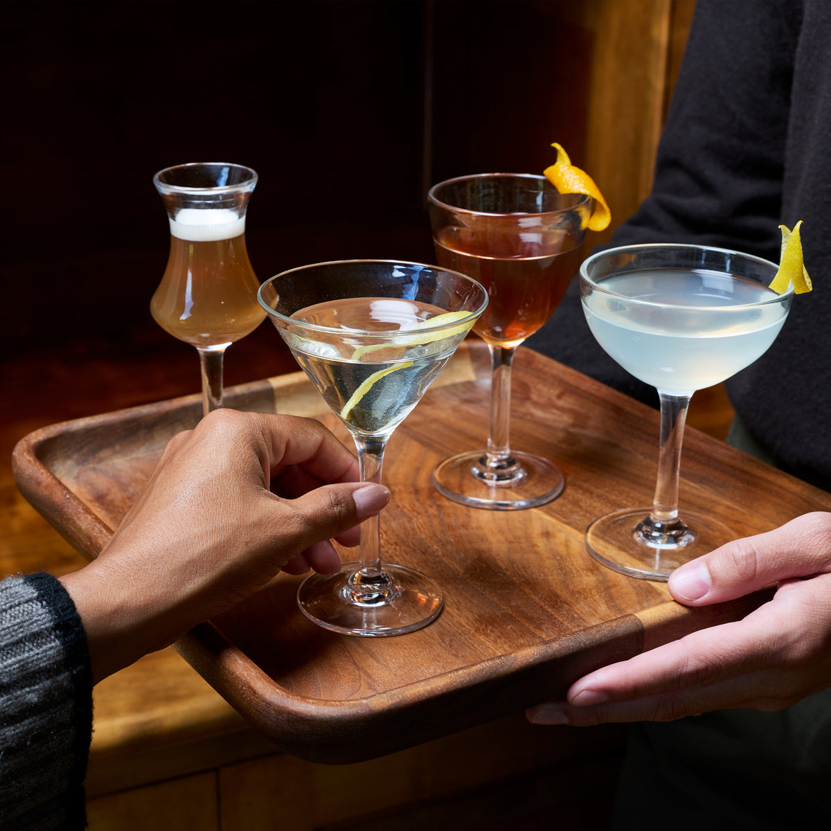 Four glass cocktail glasses with various beverages and lemon twists, held on a wooden tray.