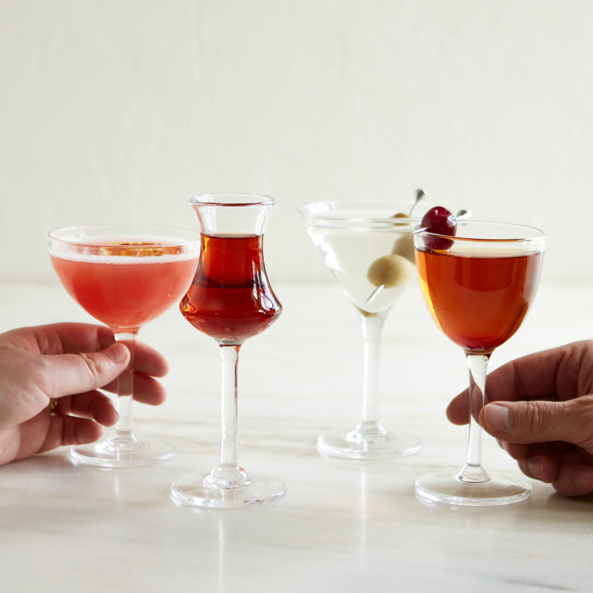 Four cocktail glasses filled with different colored beverages, two held in hands, garnished with cherries and olives.