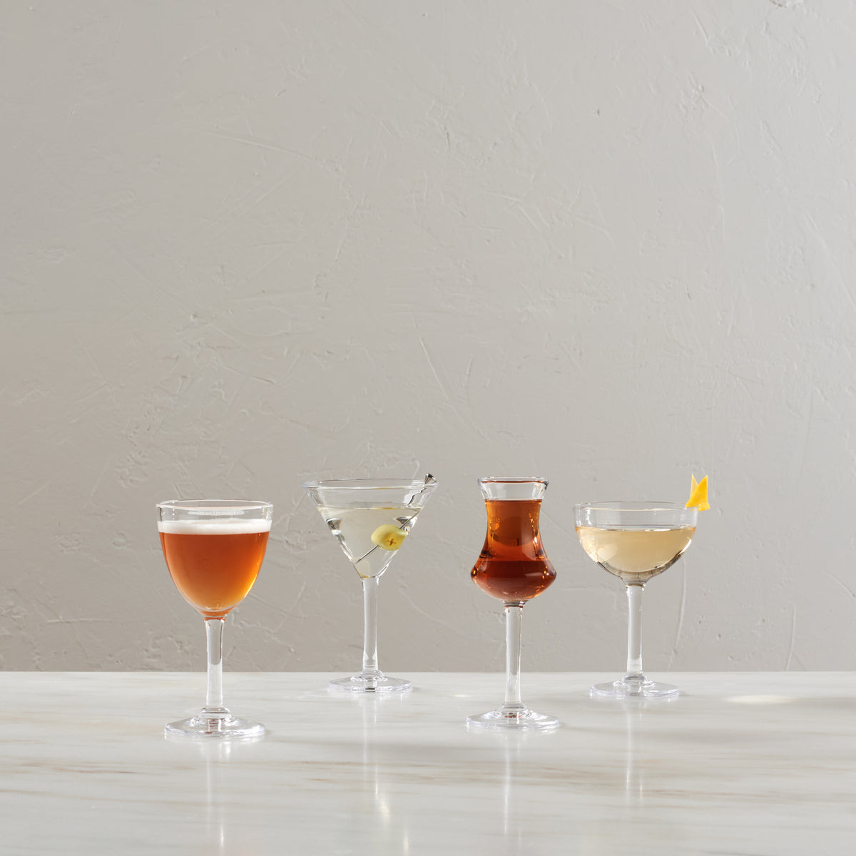 Four clear bar glasses filled with different colored cocktails, displayed on a white surface against a gray background.