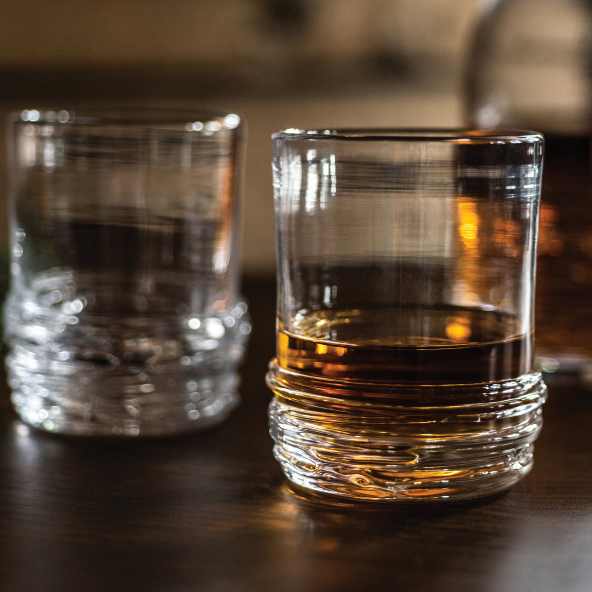 Two crystal rocks glasses with textured bases, one containing amber whiskey and one empty, on a dark surface.