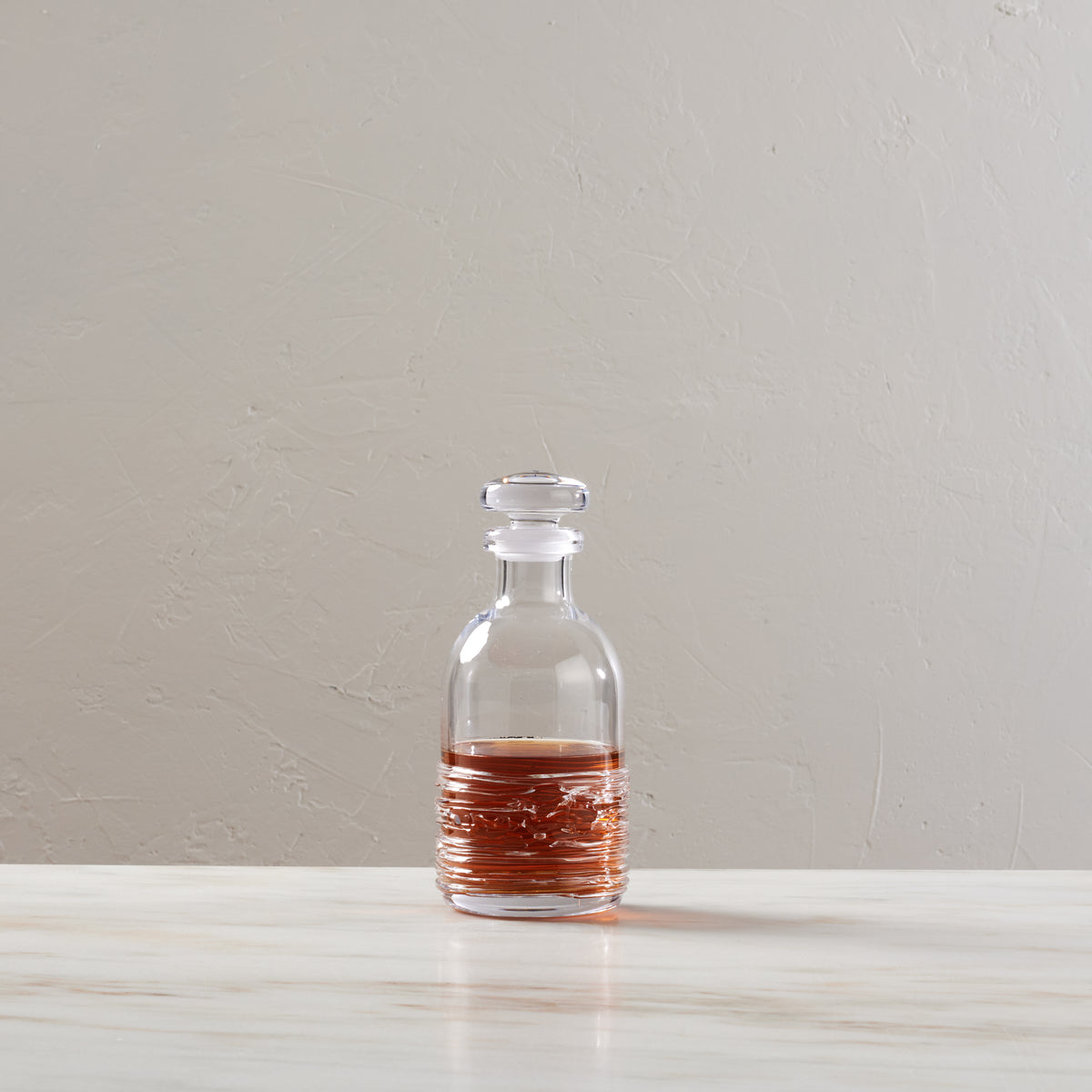 Clear glass decanter with stopper containing amber liquid, displayed on white surface against neutral background.