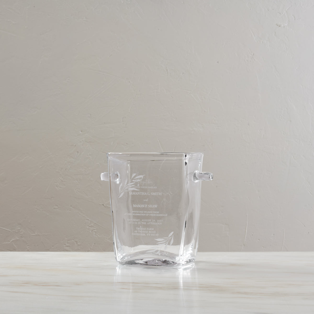 Clear glass ice bucket with two handles and engraved wedding invitation text and floral design on front.