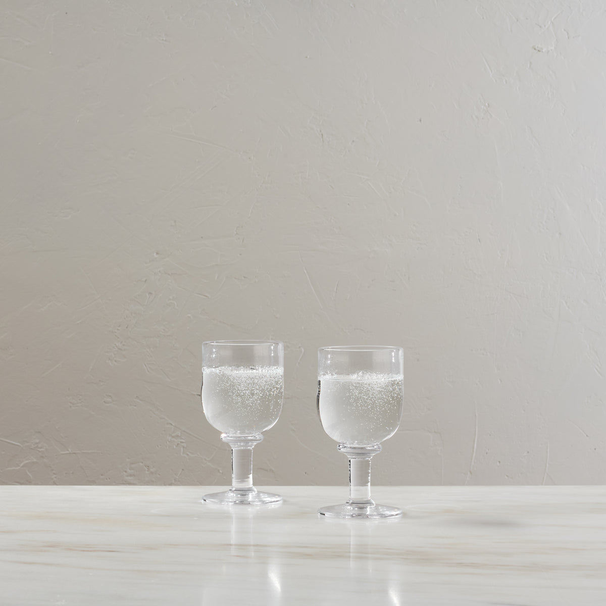 Two clear water glasses with stems, each filled with sparkling water, displayed on a white surface against a neutral backgrou
