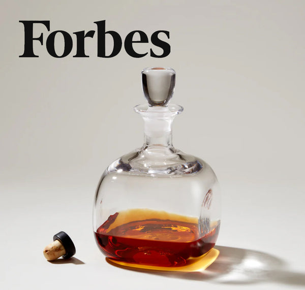 Forbes feature: Simon Pearce handblown glass decanter with a glass stopper, resting on its side with amber liquid pooled inside, on a neutral background