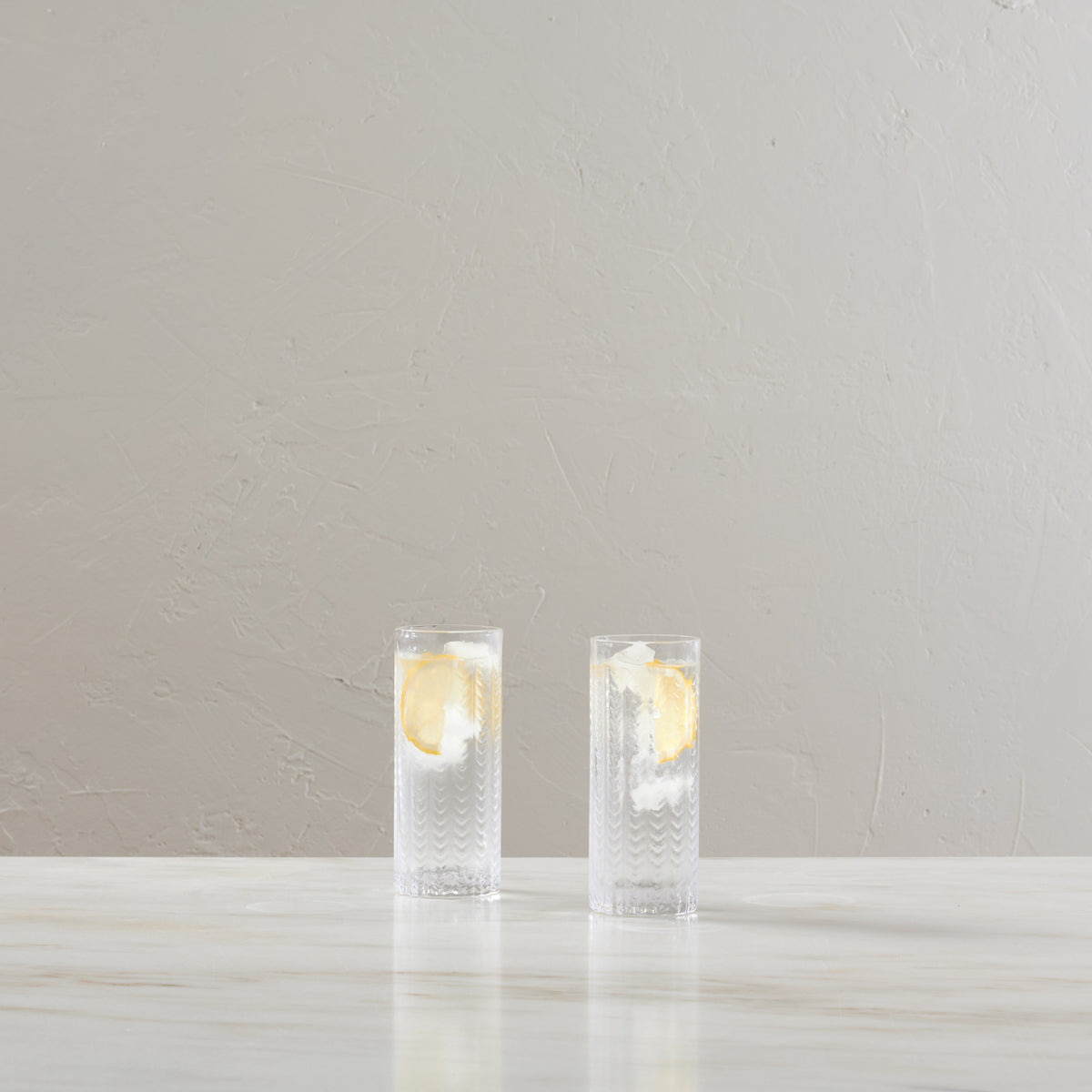 Two tall Collins glasses with textured pattern, each containing ice and lemon slices in clear liquid.