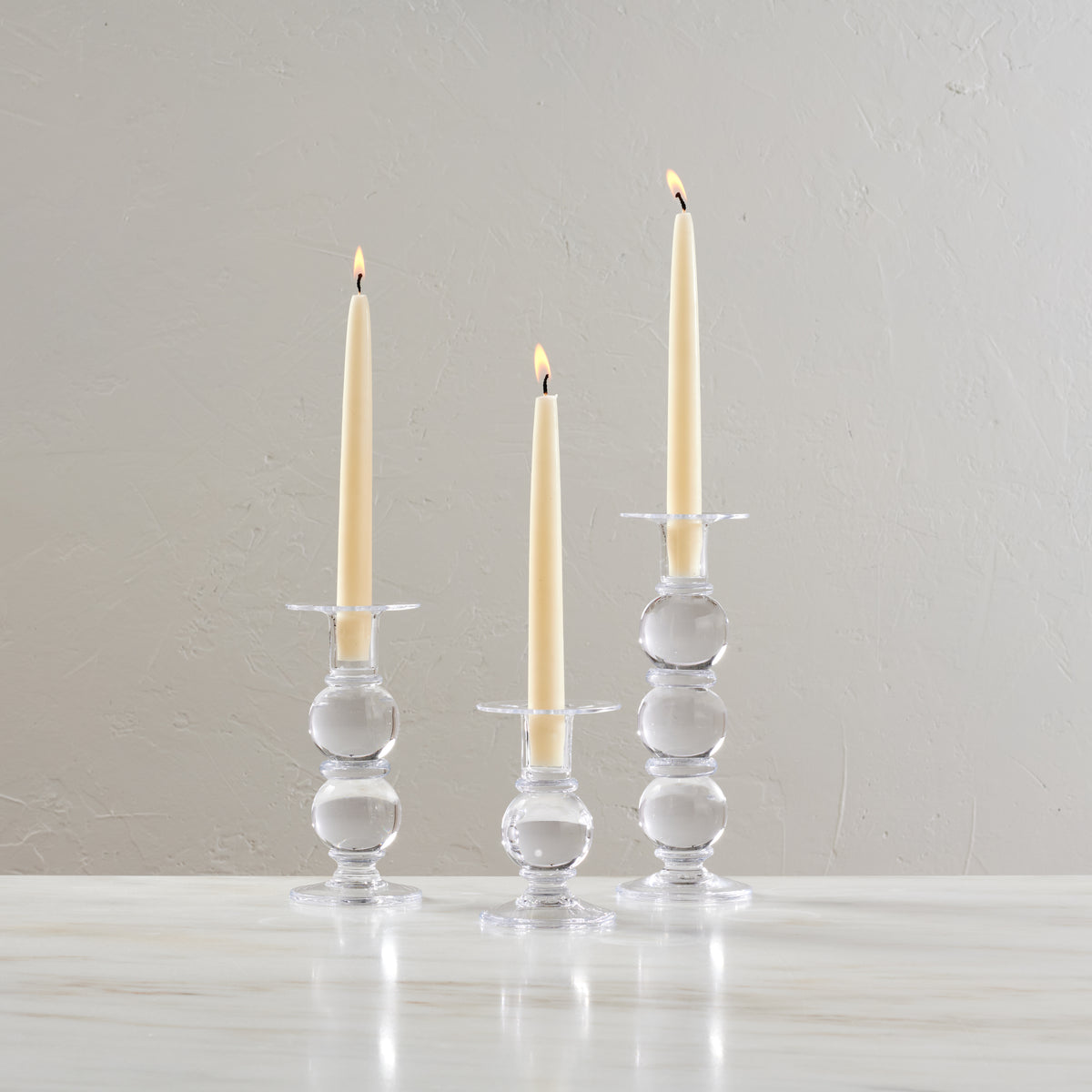 Set of three clear glass candlesticks with stacked spherical bases holding lit cream taper candles.