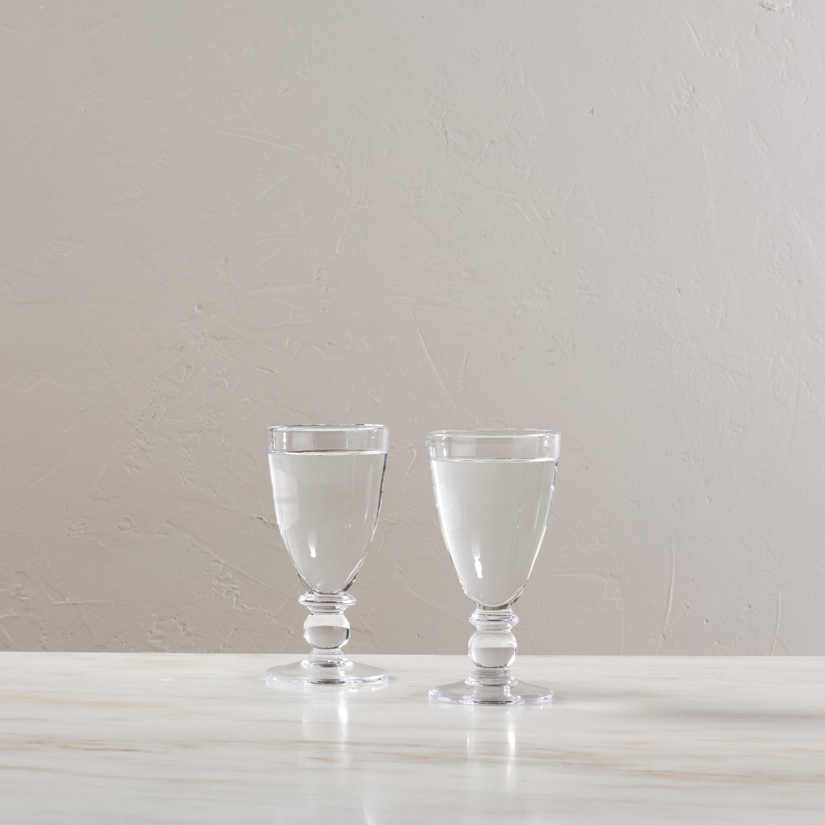 Two clear glass water glasses with pedestal bases filled with water, displayed side by side.