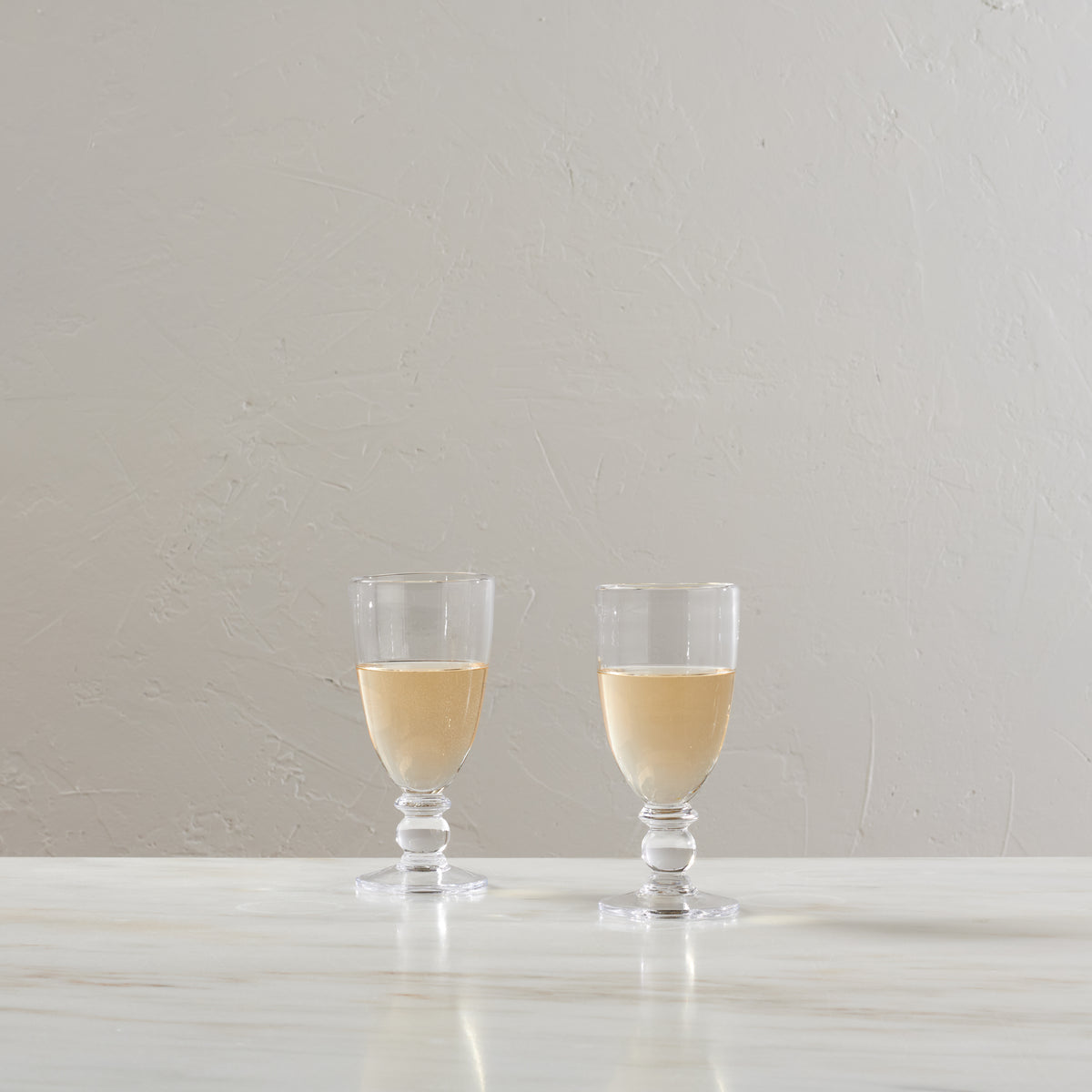 Two stemmed glasses filled with pale yellow wine on a white surface against a beige textured wall.
