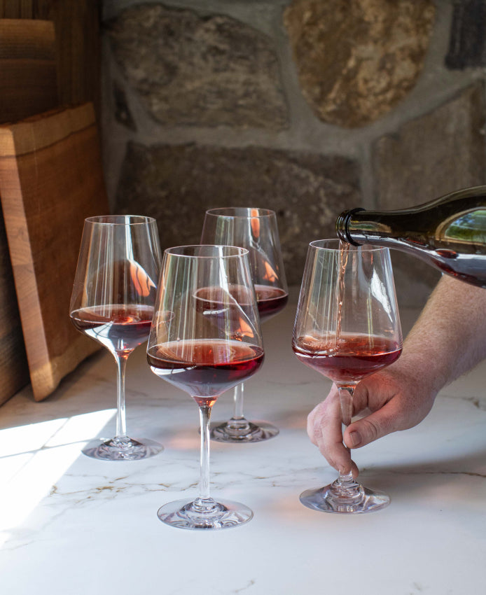 Red wine being poured from a bottle into a Simon Pearce handblown wine glass, with three additional filled wine glasses on a marble surface in front of a stone wall