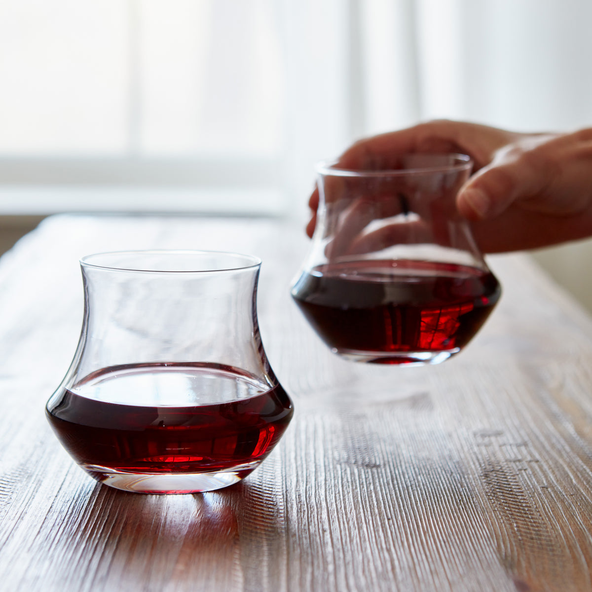 Two clear stemless wine glasses, one filled with red wine and one being held with red wine, on a wooden table.